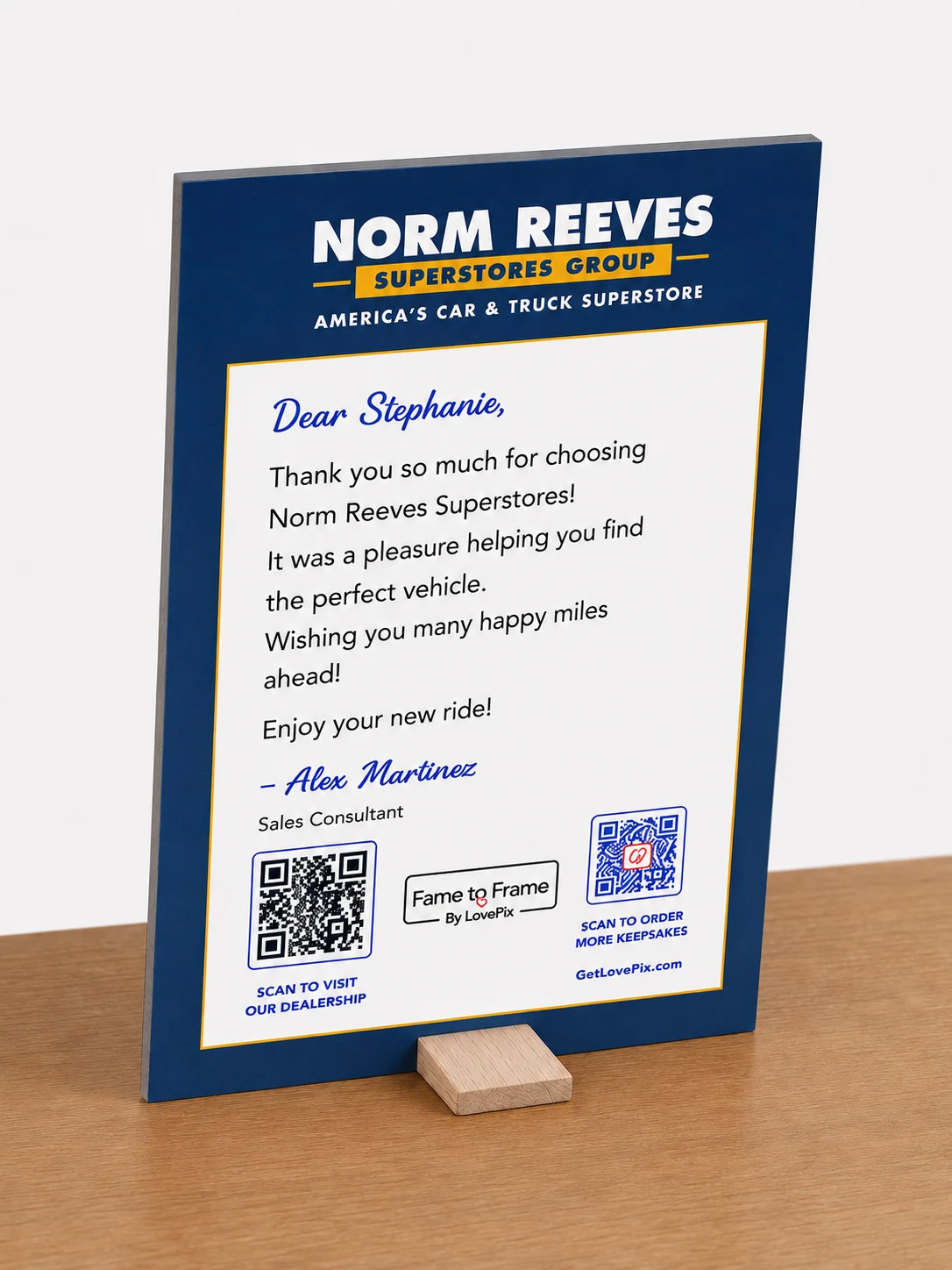 Approved rear card-back view of a Fame to Frame keepsake leaning back on its wooden support peg, with a handwritten personalized thank-you message, a larger dealership QR code, and a smaller GetLovePix.com QR code
